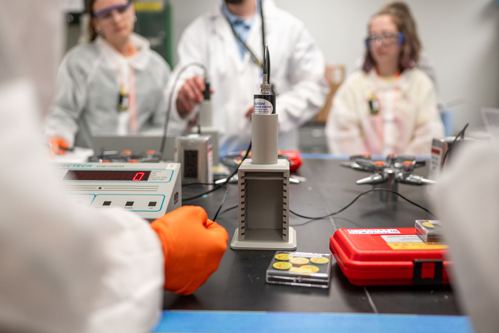 NCSS students in lab coats and safety glasses work with a radiation detector in the lab